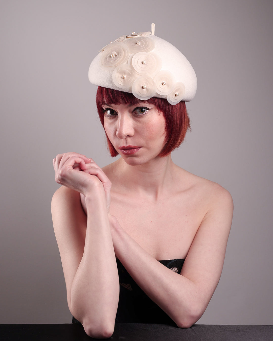The model is showcasing the top left angle of the Heaven pillbox beret hat, crafted in soft white felt and adorned with swirling ivory appliqué rosettes of different sizes and layers, each hand-finished with a delicate pearl. Designed by Merve Bayindir.