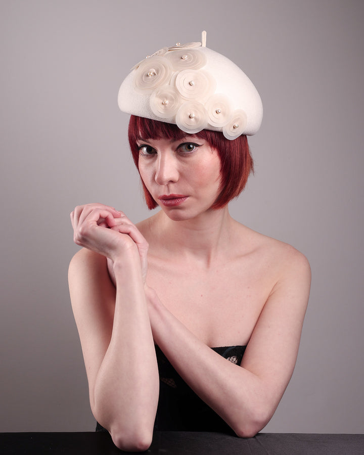 The model is showcasing the top left angle of the Heaven pillbox beret hat, crafted in soft white felt and adorned with swirling ivory appliqué rosettes of different sizes and layers, each hand-finished with a delicate pearl. Designed by Merve Bayindir.