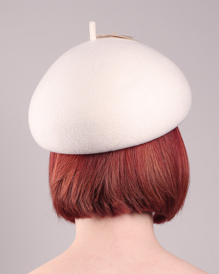 The model is showcasing the back of the Heaven pillbox beret hat, crafted in soft white felt and adorned with swirling ivory appliqué rosettes of different sizes and layers, each hand-finished with a delicate pearl. Designed by Merve Bayindir.