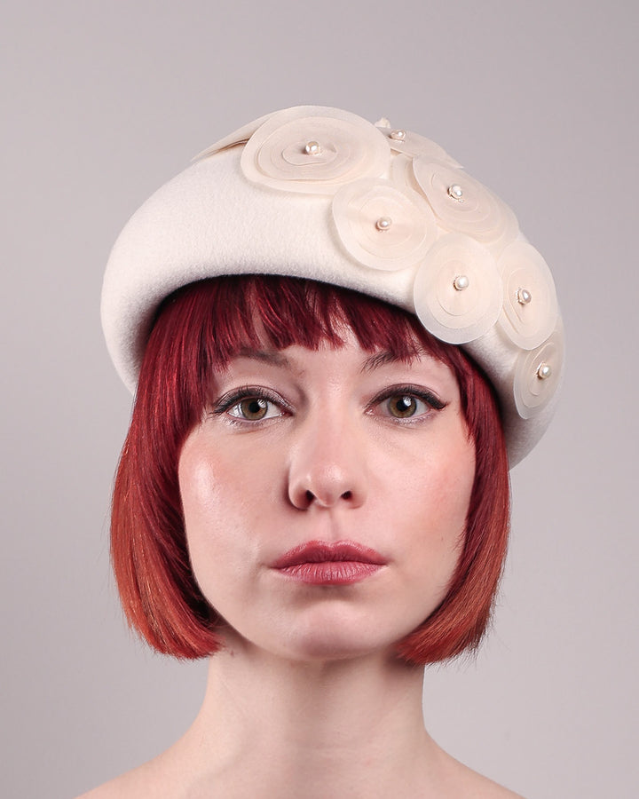The model is showcasing the front close up of the Heaven pillbox beret hat, crafted in soft white felt and adorned with swirling ivory appliqué rosettes of different sizes and layers, each hand-finished with a delicate pearl. Designed by Merve Bayindir.