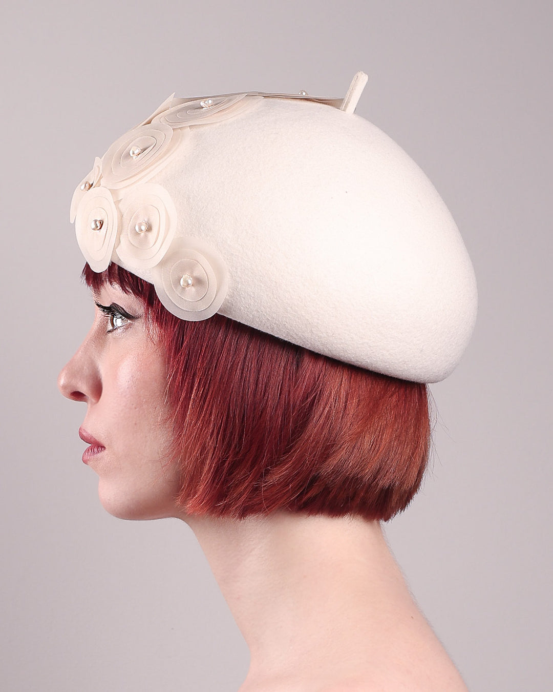 The model is showcasing the left side close up of the Heaven pillbox beret hat, crafted in soft white felt and adorned with swirling ivory appliqué rosettes of different sizes and layers, each hand-finished with a delicate pearl. Designed by Merve Bayindir.