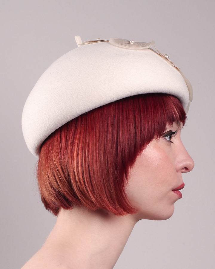 The model is showcasing the right side of the Heaven pillbox beret hat, crafted in soft white felt and adorned with swirling ivory appliqué rosettes of different sizes and layers, each hand-finished with a delicate pearl. Designed by Merve Bayindir.