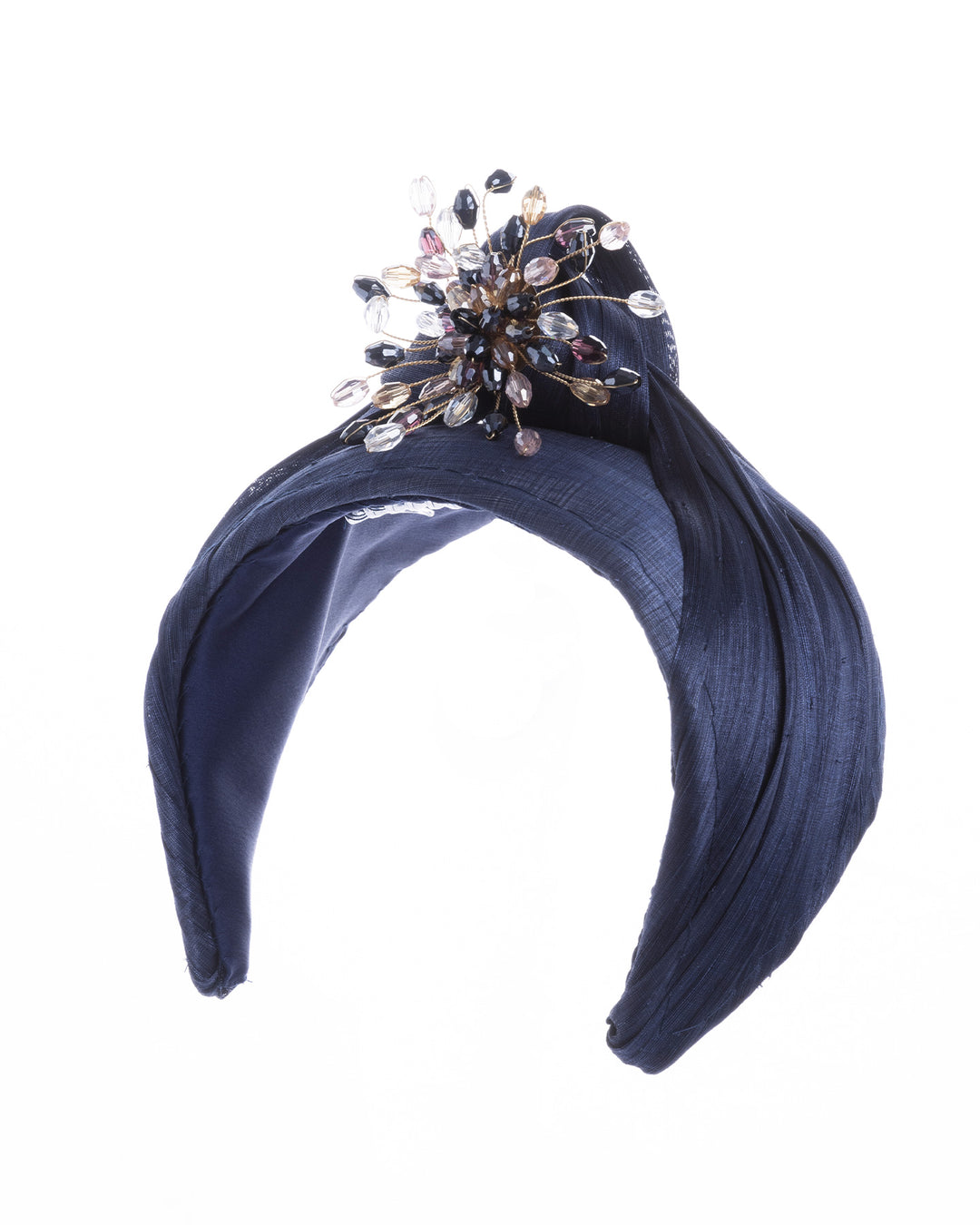 Navy blue structured half-hat in textured fabric, featuring a sculptural twisted silhouette and embellished with a statement crystal burst brooch in gold, smoky, and blush tones. Designed and made by Merve Bayindir Millinery.
