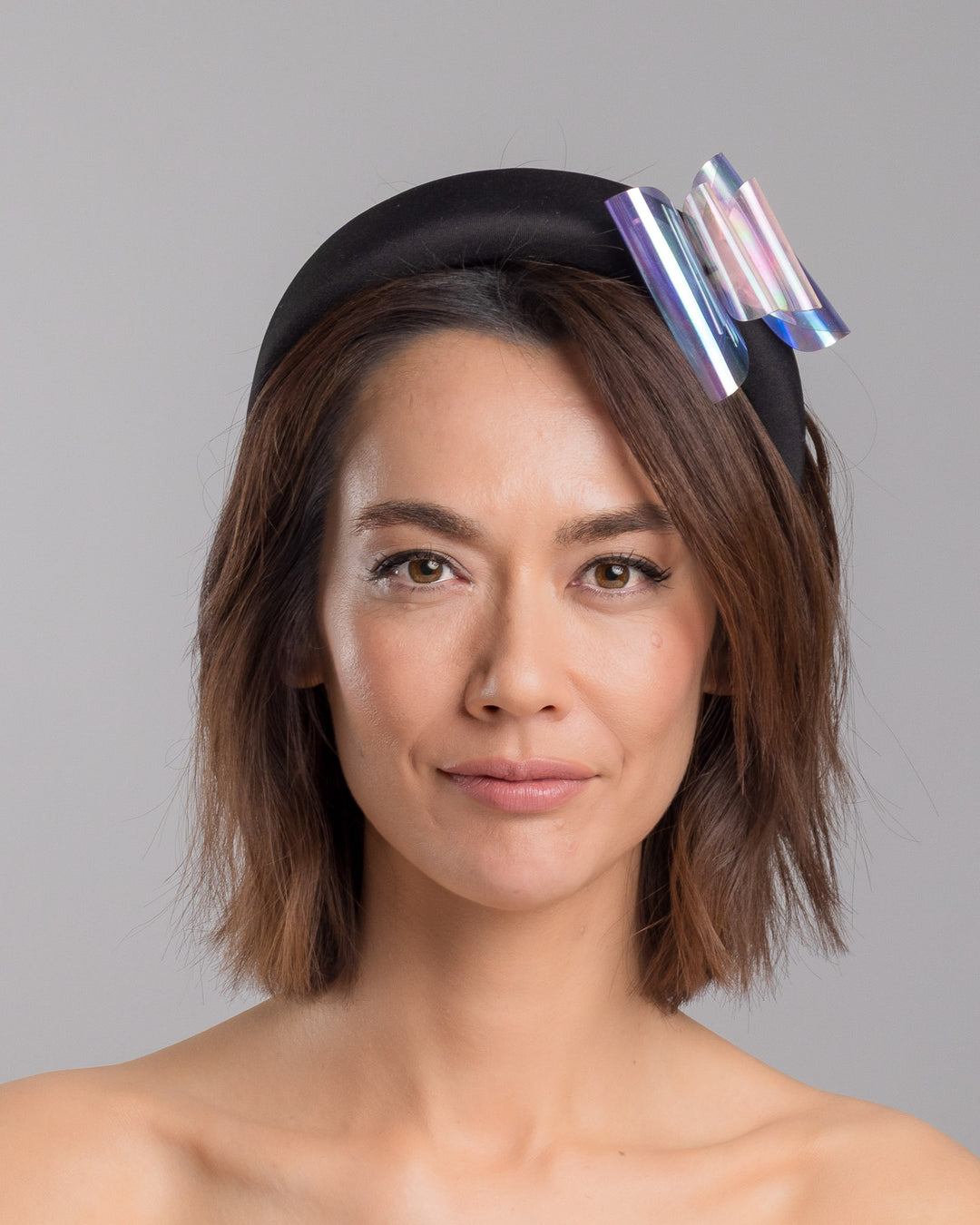 The model is showcasing the front side close up of the black padded headband featuring an oversized sculptural bow made from iridescent translucent panels with a holographic sheen, accented at the center with a soft pink geometric embellishment. Designed and made by Merve Bayindir Millinery.
