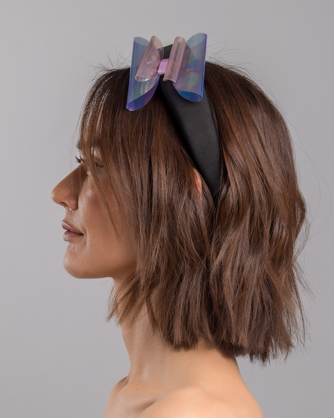 The model is showcasing the left side close up of the black padded headband featuring an oversized sculptural bow made from iridescent translucent panels with a holographic sheen, accented at the center with a soft pink geometric embellishment. Designed and made by Merve Bayindir Millinery.
