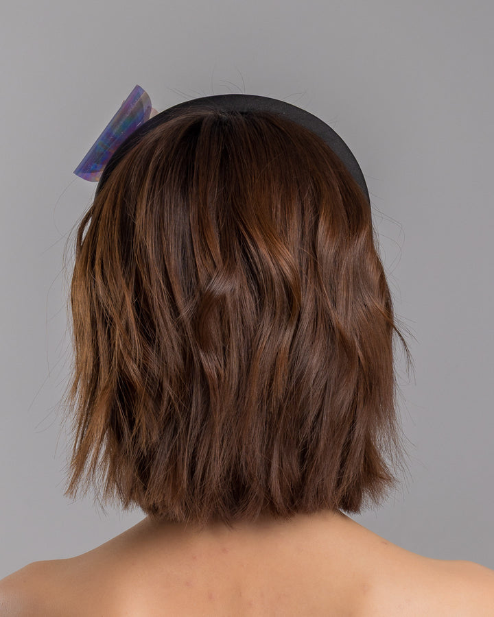 The model is showcasing the back side close up of the black padded headband featuring an oversized sculptural bow made from iridescent translucent panels with a holographic sheen, accented at the center with a soft pink geometric embellishment. Designed and made by Merve Bayindir Millinery.
