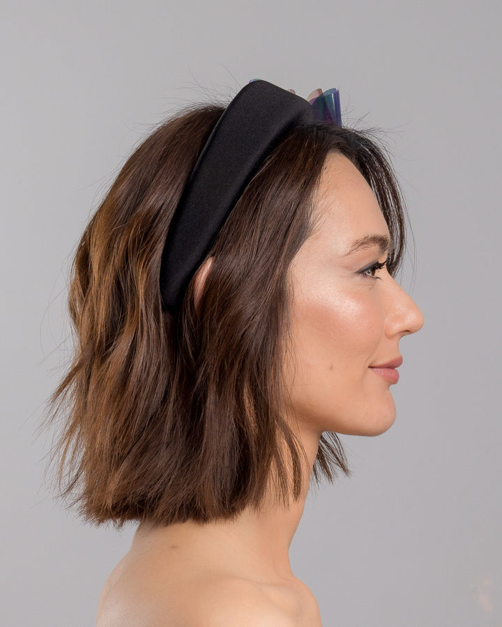 The model is showcasing the right side close up of the black padded headband featuring an oversized sculptural bow made from iridescent translucent panels with a holographic sheen, accented at the center with a soft pink geometric embellishment. Designed and made by Merve Bayindir Millinery.
