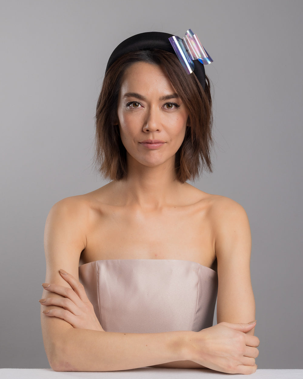 The female model is posing, wearing 
Irix, the black padded headband featuring an oversized sculptural bow made from iridescent translucent panels with a holographic sheen, accented at the center with a soft pink geometric embellishment. Designed and made by Merve Bayindir Millinery.