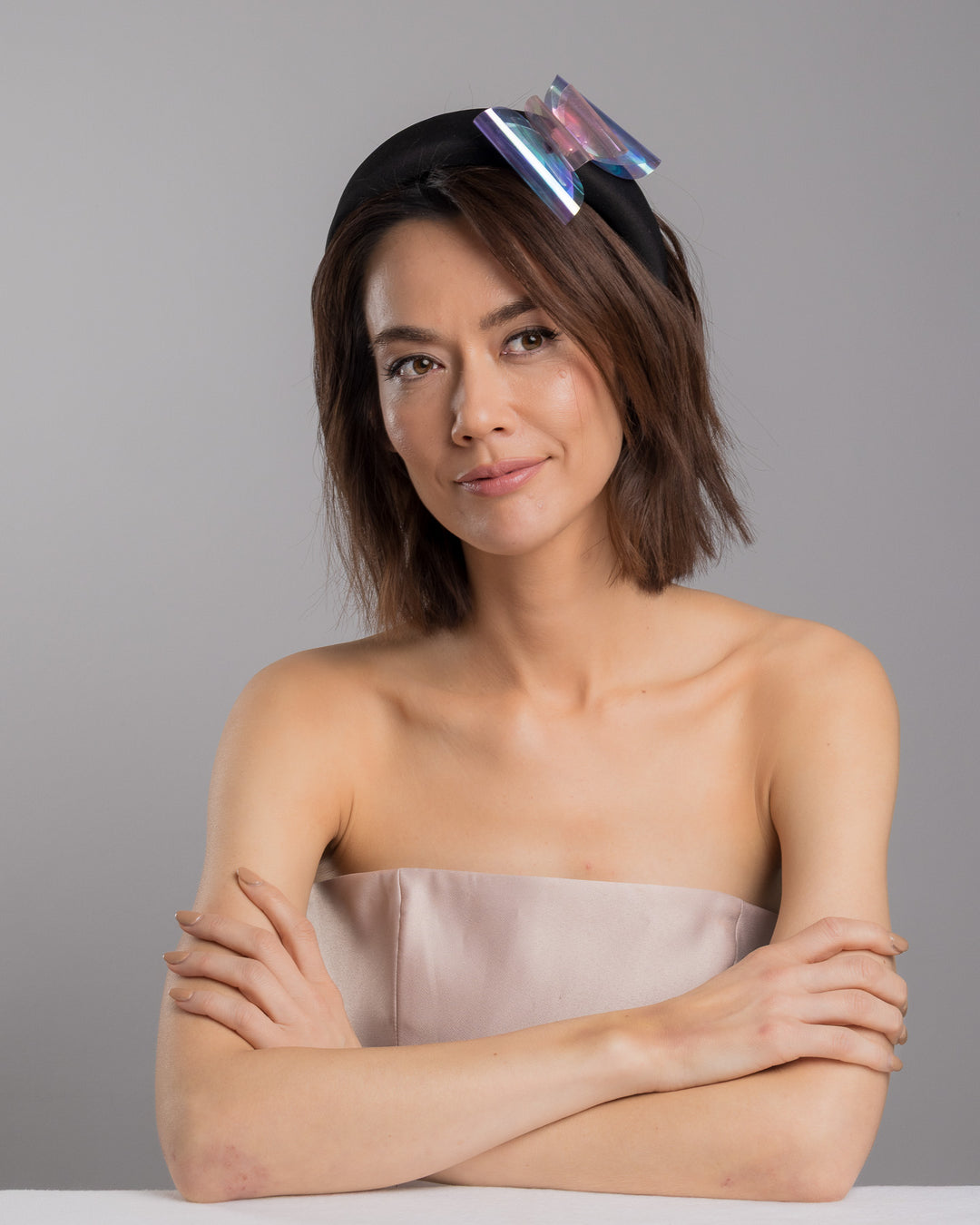 The female model is posing, wearing 
Irix, the black padded headband featuring an oversized sculptural bow made from iridescent translucent panels with a holographic sheen, accented at the center with a soft pink geometric embellishment. Designed and made by Merve Bayindir Millinery.
