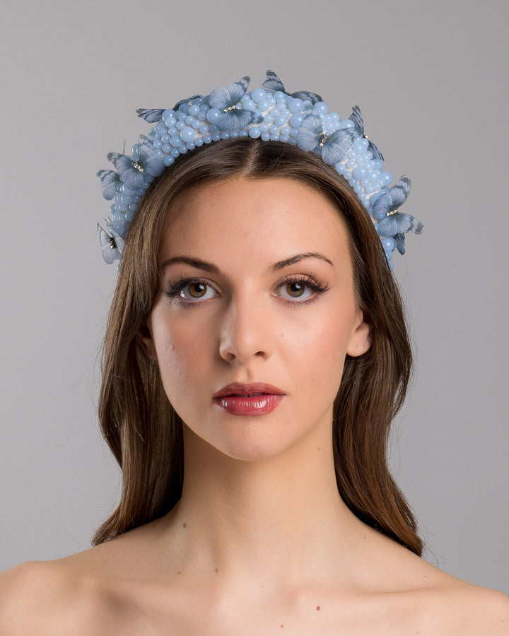 Model is showcasing the front side close up of high headband decorated with different sized mat blue beads and scattered blue organza butterflies with crystal centres. Made by Merve Bayindir millinery.