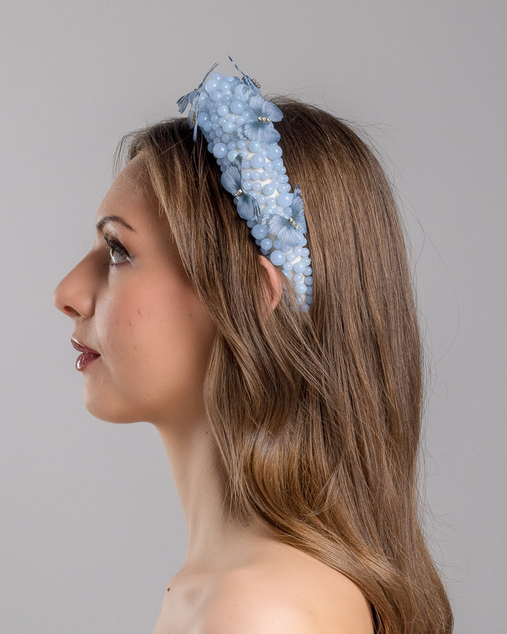 Model is showcasing the left side close up of high headband decorated with different sized mat blue beads and scattered blue organza butterflies with crystal centres. Made by Merve Bayindir millinery.