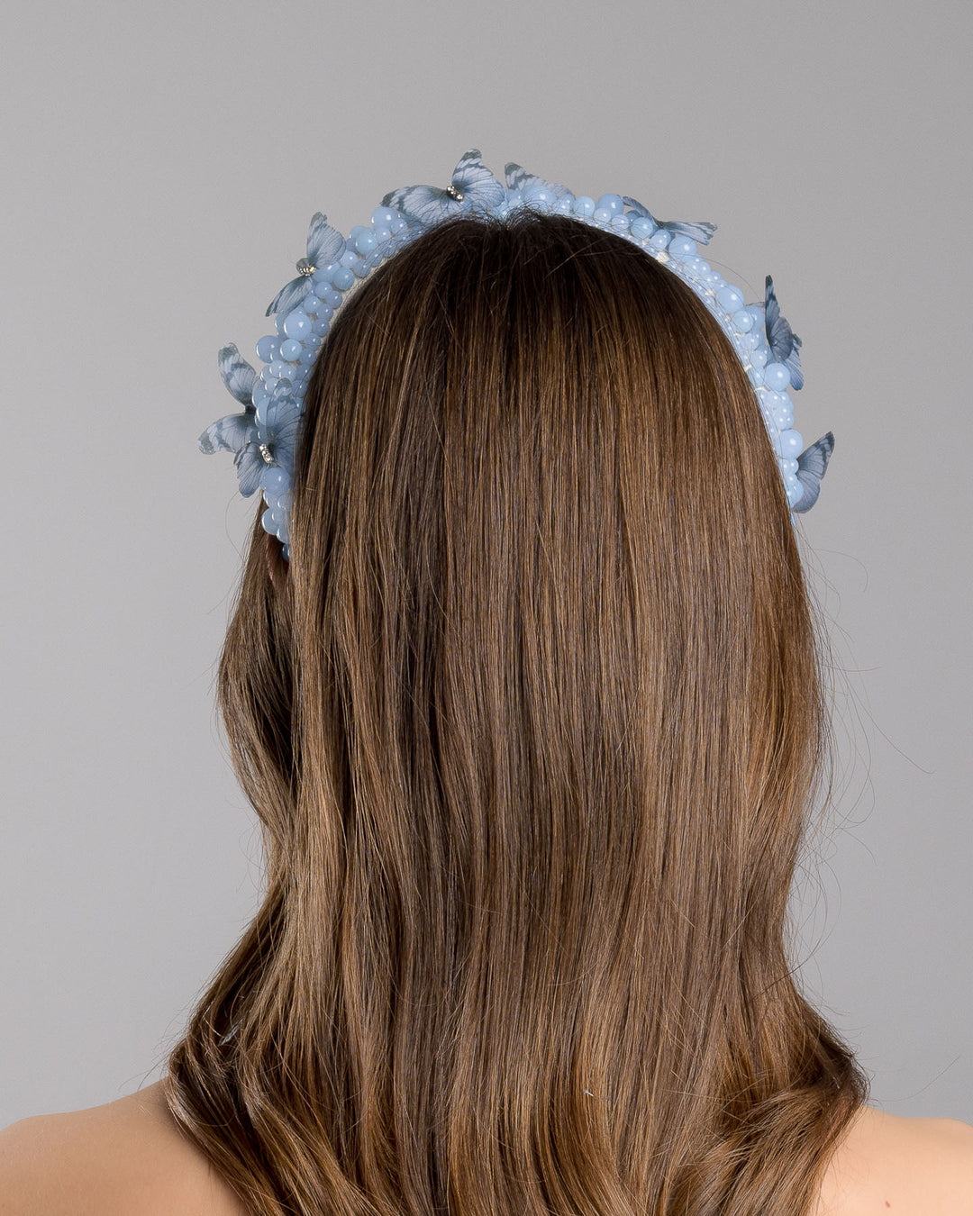 Model is showcasing the back side close up of high headband decorated with different sized mat blue beads and scattered blue organza butterflies with crystal centres. Made by Merve Bayindir millinery.