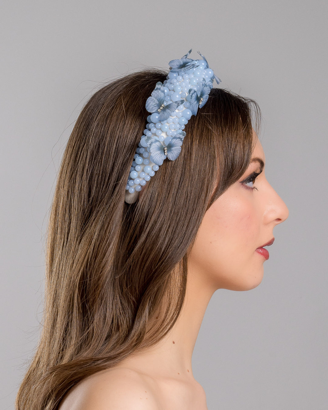 Model is showcasing the right side close up of high headband decorated with different sized mat blue beads and scattered blue organza butterflies with crystal centres. Made by Merve Bayindir millinery.