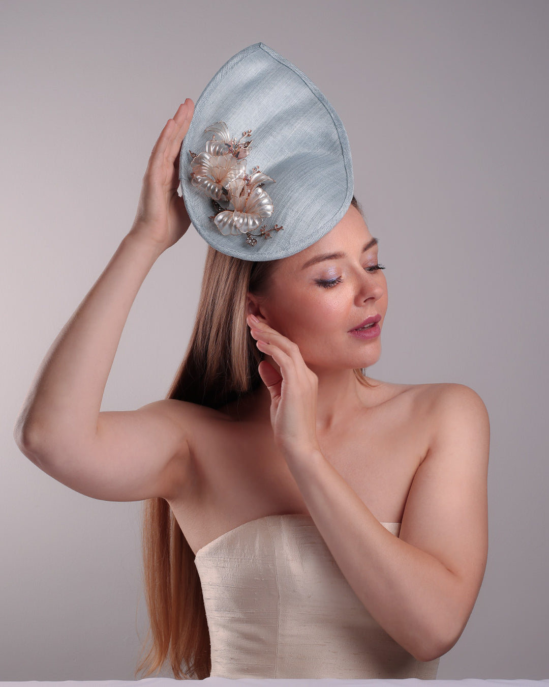 The model is showcasing the front of the blue fascinator from the angle. A blue tall fascinator resembling shell like curves, decorated with a see through resin flowers. Simple yet elegant fascinator is fitted with a wire headband and elastic