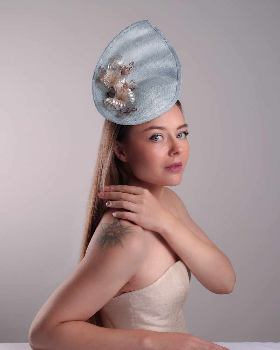 The model is showcasing the front of the blue fascinator from the right angle. A blue tall fascinator resembling shell like curves, decorated with a see through resin flowers. Simple yet elegant fascinator is fitted with a wire headband and elastic