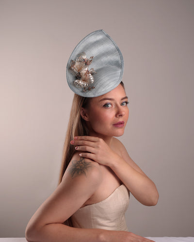 The model is showcasing the front of the blue fascinator from the right angle. A blue tall fascinator resembling shell like curves, decorated with a see through resin flowers. Simple yet elegant fascinator is fitted with a wire headband and elastic