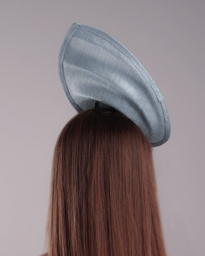 The model is showcasing the back of a blue tall fascinator resembling shell like curves, decorated with a see through resin flowers. Simple yet elegant fascinator is fitted with a wire headband and elastic