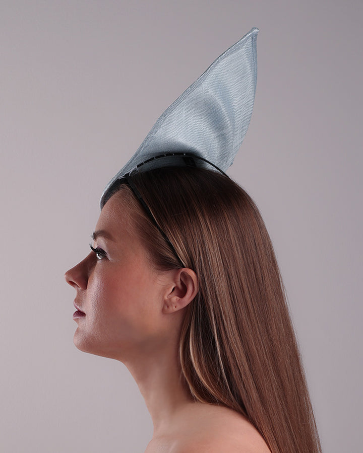 The model is showcasing the left side of a blue tall fascinator resembling shell like curves, decorated with a see through resin flowers. Simple yet elegant fascinator is fitted with a wire headband and elastic