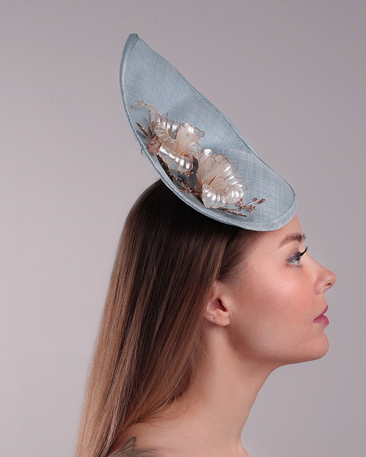 The model is showcasing the right side of a blue tall fascinator resembling shell like curves, decorated with a see through resin flowers. Simple yet elegant fascinator is fitted with a wire headband and elastic