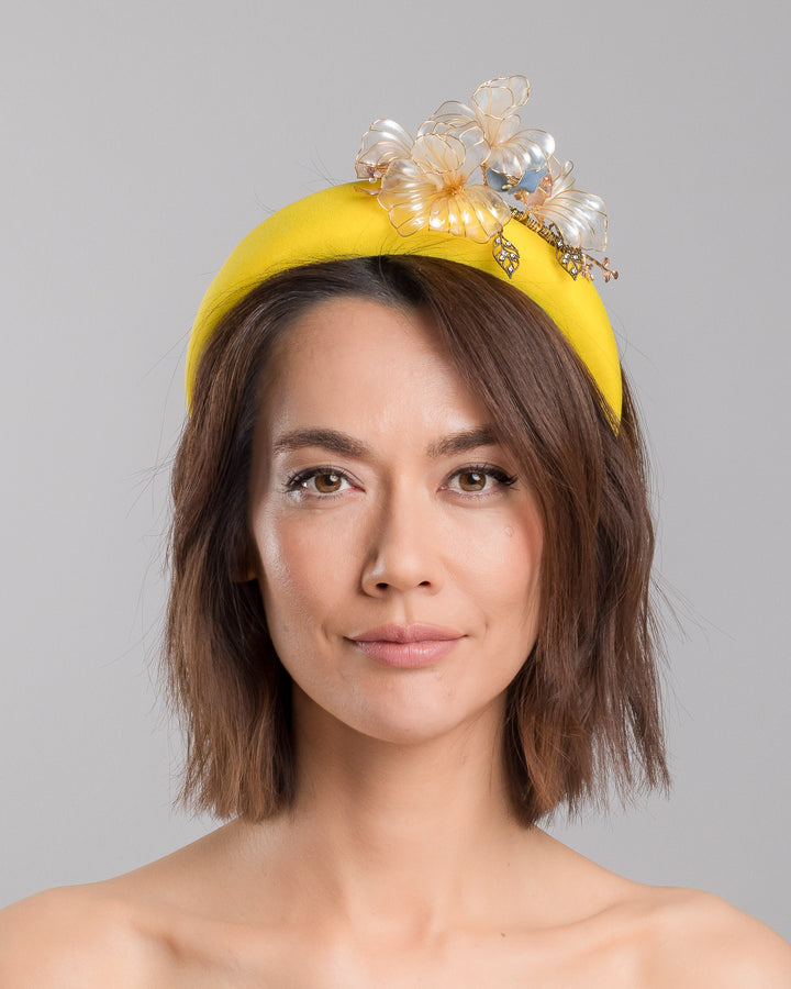 Model is showcasing the front of high yellow headband decorated with wired sheer resin flowers and small metal adornments. Made by Merve Bayindir. 