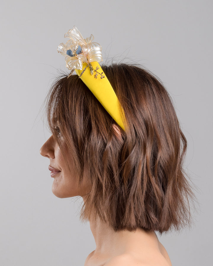 Model is showcasing the left side of high yellow headband decorated with wired sheer resin flowers and small metal adornments. Made by Merve Bayindir. 