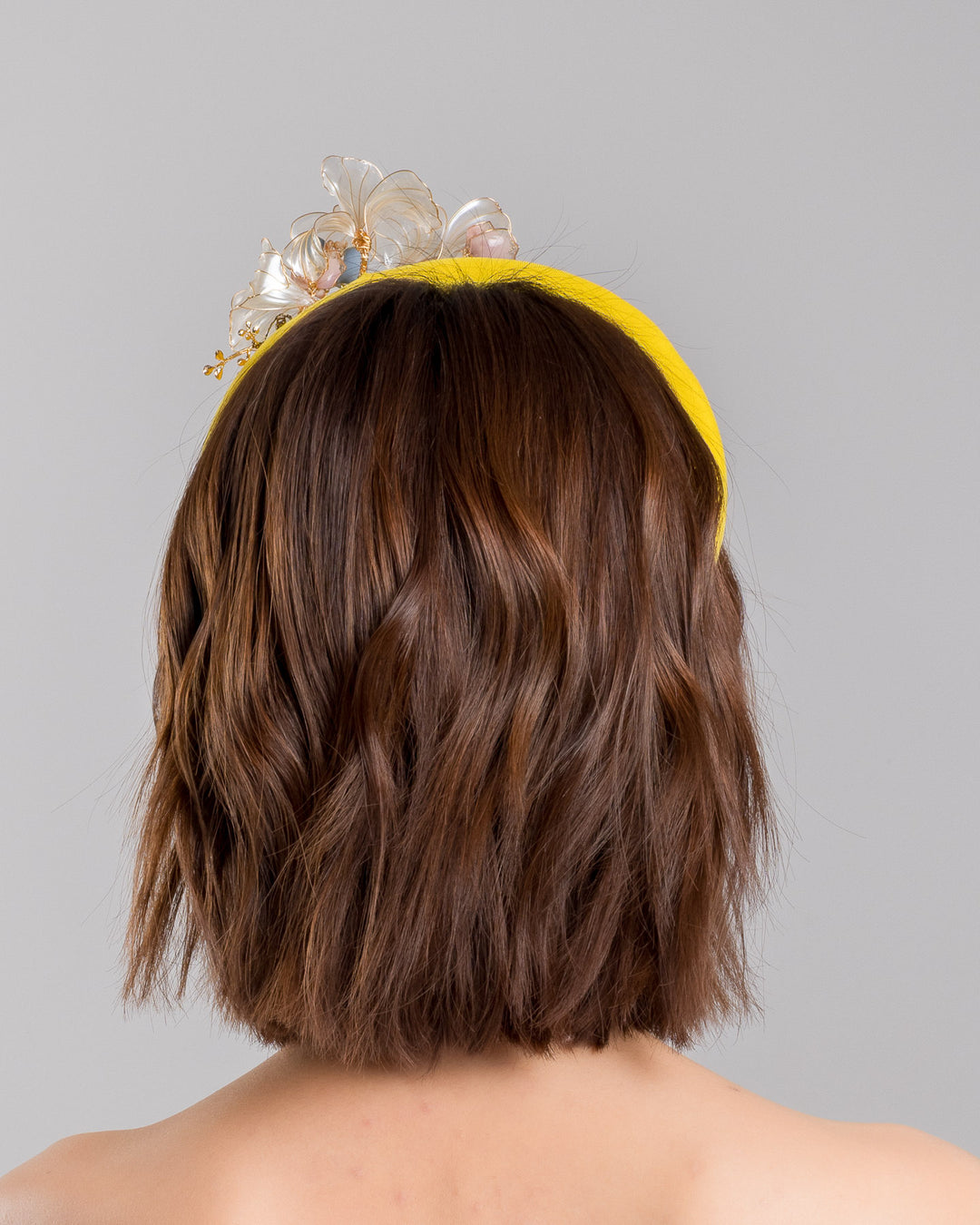 Model is showcasing the back of high yellow headband decorated with wired sheer resin flowers and small metal adornments. Made by Merve Bayindir. 