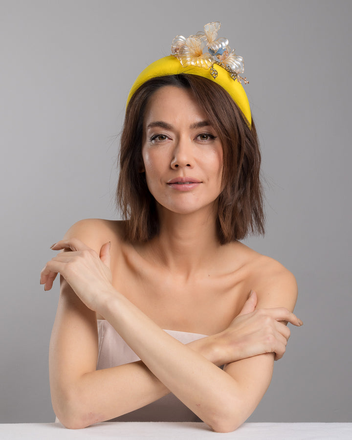 Model is posing wearing Illecebra high yellow headband decorated with wired sheer resin flowers and small metal adornments. Made by Merve Bayindir Millinery.