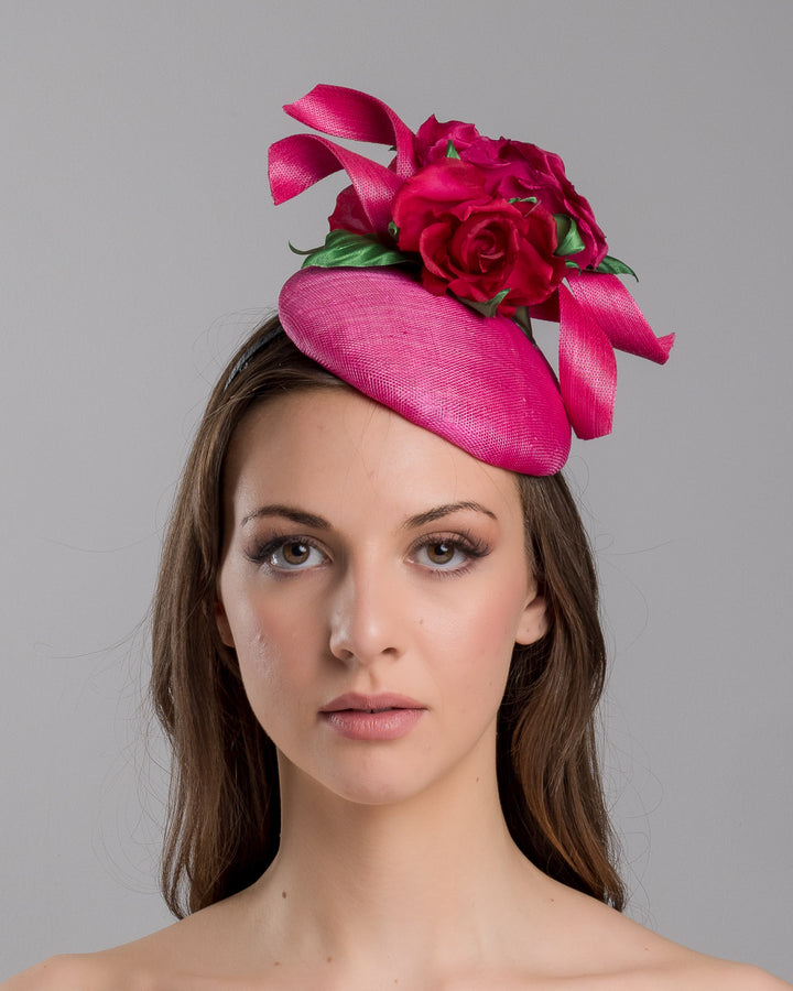 The model is showcasing the front side close up of the hot pink sinamay fascinator with a sculpted asymmetrical base, topped with deep red rose blooms and green leaves, mounted on a slim black headband. Designed and made by Merve Bayindir Millinery.
