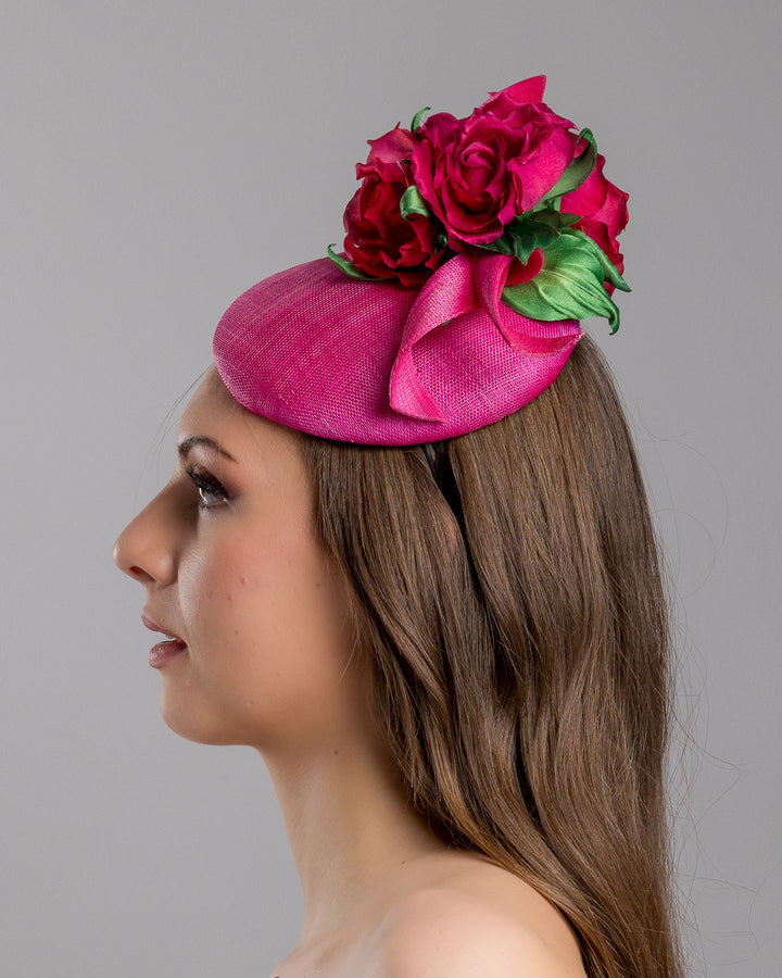 The model is showcasing the left  side close up of the hot pink sinamay fascinator with a sculpted asymmetrical base, topped with deep red rose blooms and green leaves, mounted on a slim black headband. Designed and made by Merve Bayindir Millinery.
