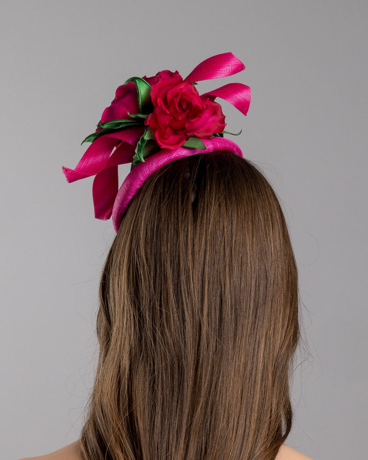 The model is showcasing the back side close up of the hot pink sinamay fascinator with a sculpted asymmetrical base, topped with deep red rose blooms and green leaves, mounted on a slim black headband. Designed and made by Merve Bayindir Millinery.
