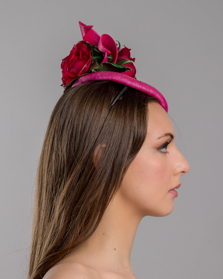 The model is showcasing the right side close up of the hot pink sinamay fascinator with a sculpted asymmetrical base, topped with deep red rose blooms and green leaves, mounted on a slim black headband. Designed and made by Merve Bayindir Millinery.

