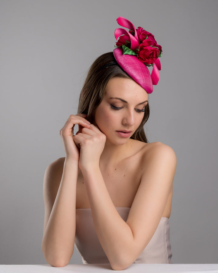 The female model is posing, wearing
Jaylin, the hot pink sinamay fascinator with a sculpted asymmetrical base, topped with deep red rose blooms and green leaves, mounted on a slim black headband. Designed and made by Merve Bayindir Millinery.
