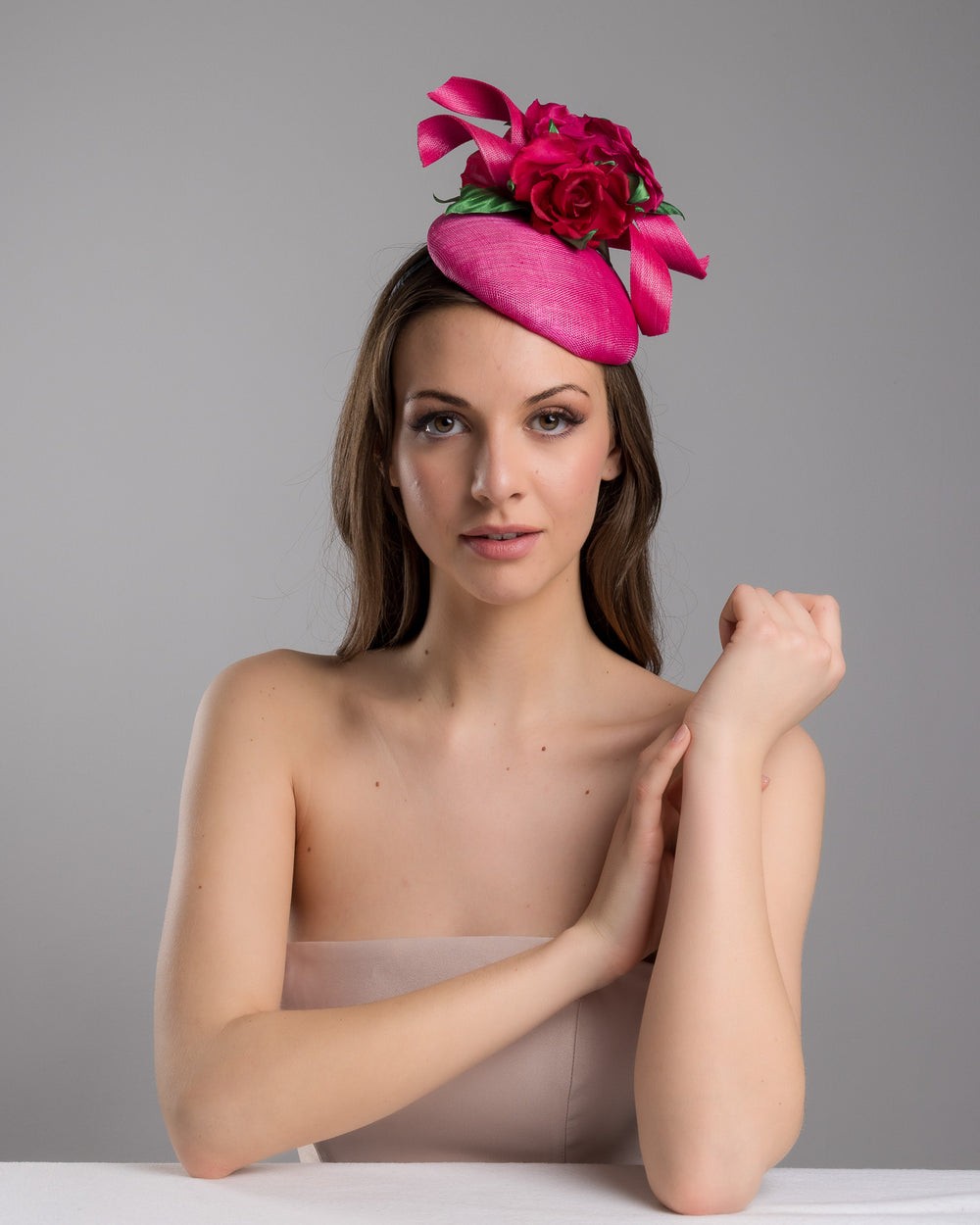 The female model is posing, wearing
Jaylin, the hot pink sinamay fascinator with a sculpted asymmetrical base, topped with deep red rose blooms and green leaves, mounted on a slim black headband. Designed and made by Merve Bayindir Millinery.
