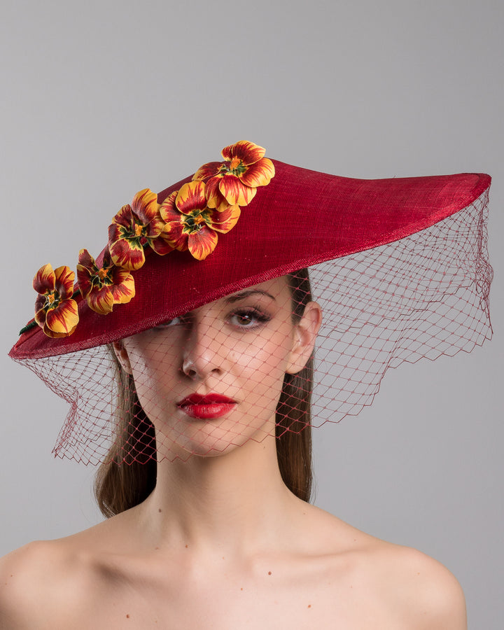 The model is showcasing the front side close up of the deep red sinamay wide-brim fascinator with a sculptural pointed shape, decorated with orange and burgundy pansy flowers and a matching red birdcage veil. Designed and made by Merve Bayindir Millinery.
