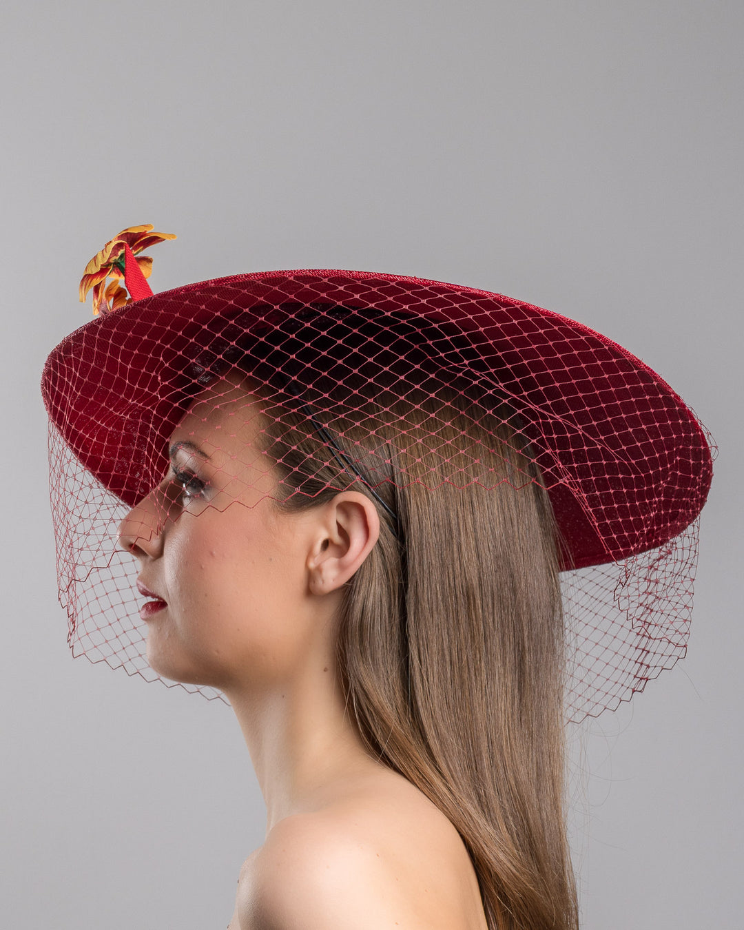 The model is showcasing the left side close up of the deep red sinamay wide-brim fascinator with a sculptural pointed shape, decorated with orange and burgundy pansy flowers and a matching red birdcage veil. Designed and made by Merve Bayindir Millinery.
