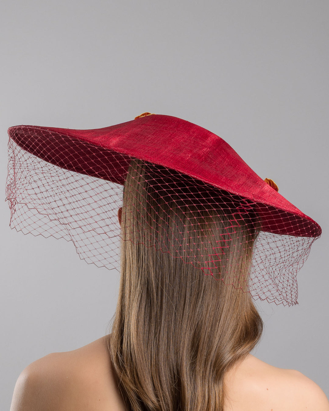 The model is showcasing the back  side close up of the deep red sinamay wide-brim fascinator with a sculptural pointed shape, decorated with orange and burgundy pansy flowers and a matching red birdcage veil. Designed and made by Merve Bayindir Millinery.
