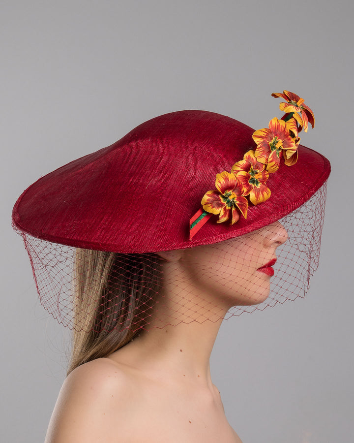 The model is showcasing the right side close up of the deep red sinamay wide-brim fascinator with a sculptural pointed shape, decorated with orange and burgundy pansy flowers and a matching red birdcage veil. Designed and made by Merve Bayindir Millinery.
