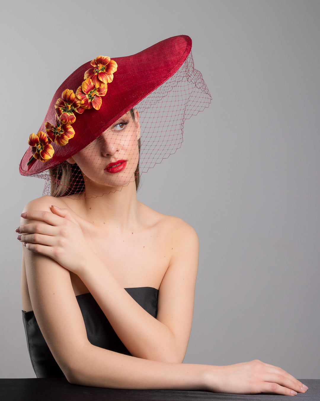 The female model is posing, wearing
Jericho, the deep red sinamay wide-brim fascinator with a sculptural pointed shape, decorated with orange and burgundy pansy flowers and a matching red birdcage veil. Designed and made by Merve Bayindir Millinery.
