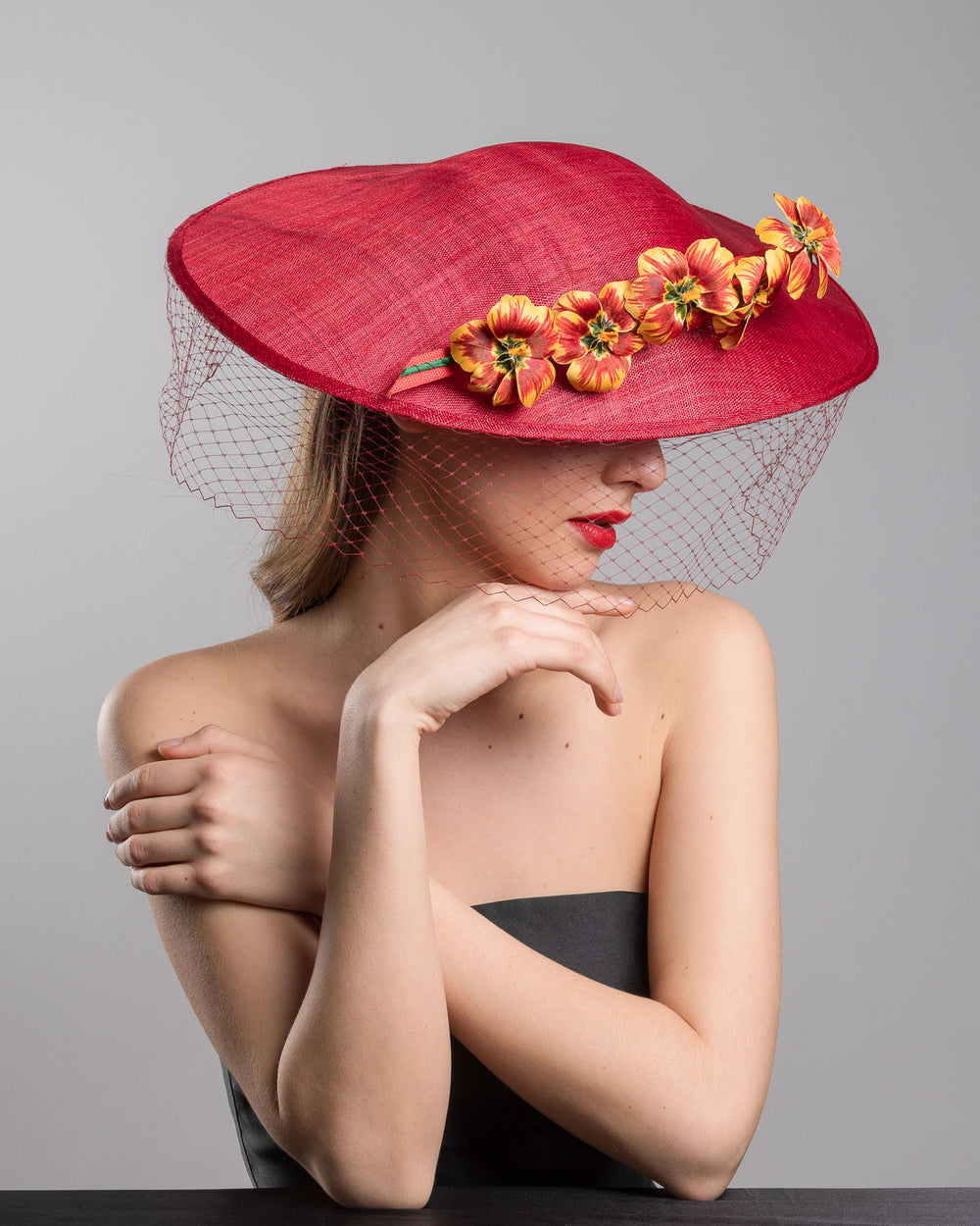 The female model is posing, wearing
Jericho, the deep red sinamay wide-brim fascinator with a sculptural pointed shape, decorated with orange and burgundy pansy flowers and a matching red birdcage veil. Designed and made by Merve Bayindir Millinery.
