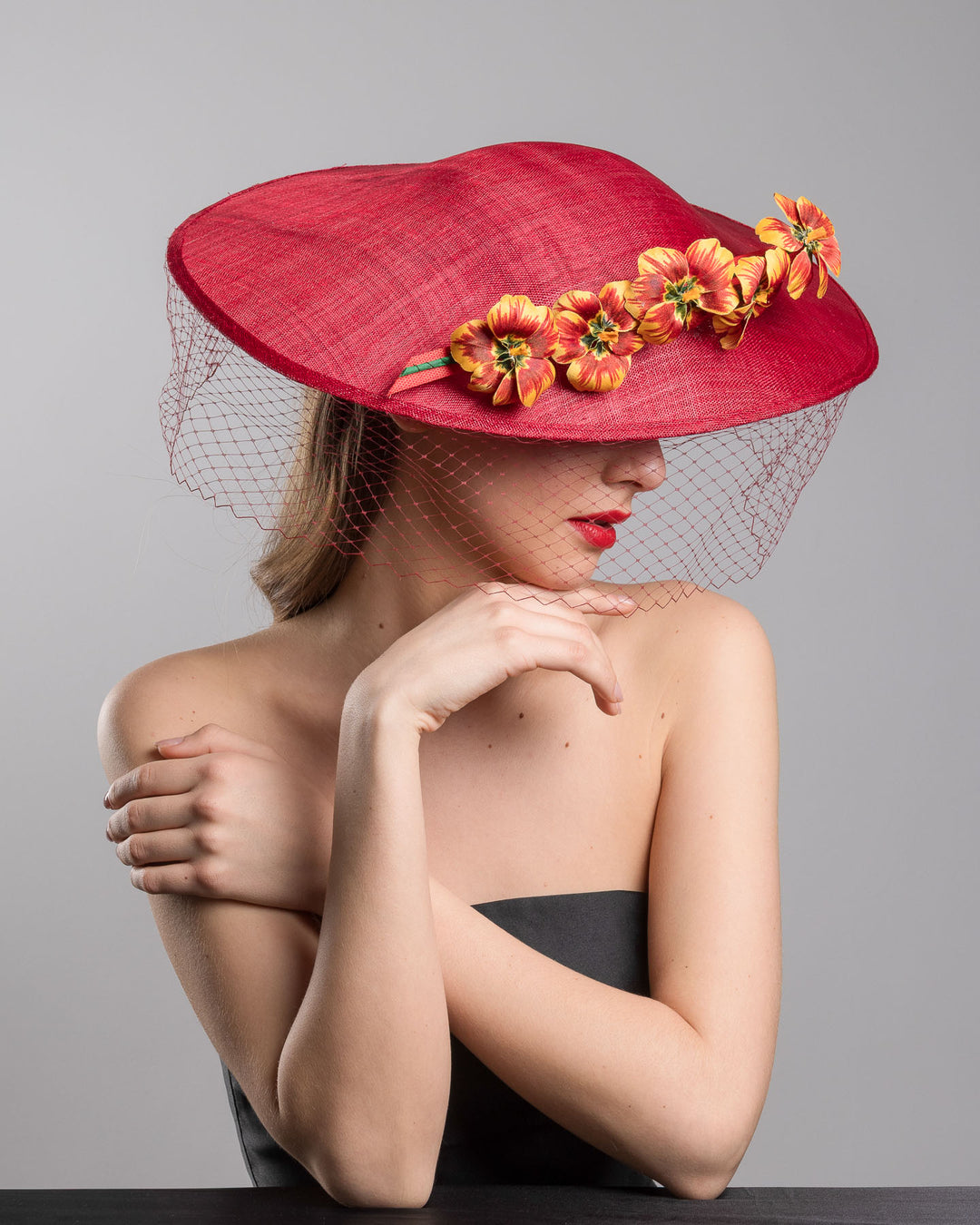 The female model is posing, wearing
Jericho, the deep red sinamay wide-brim fascinator with a sculptural pointed shape, decorated with orange and burgundy pansy flowers and a matching red birdcage veil. Designed and made by Merve Bayindir Millinery.
