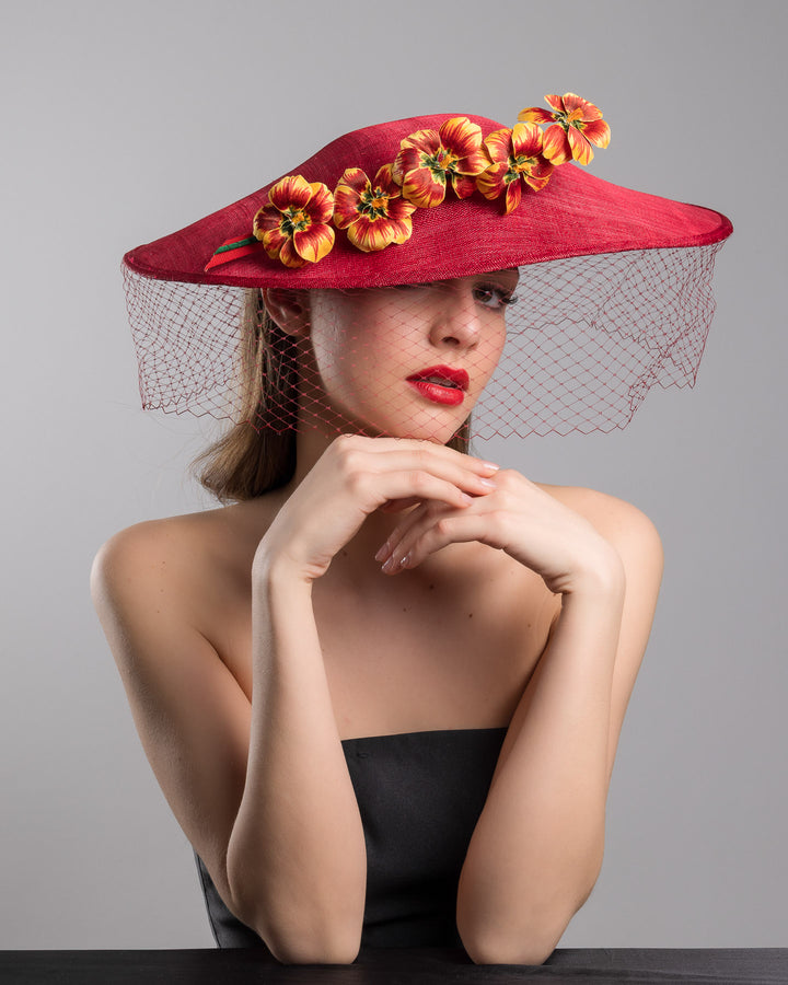 The female model is posing, wearing
Jericho, the deep red sinamay wide-brim fascinator with a sculptural pointed shape, decorated with orange and burgundy pansy flowers and a matching red birdcage veil. Designed and made by Merve Bayindir Millinery.
