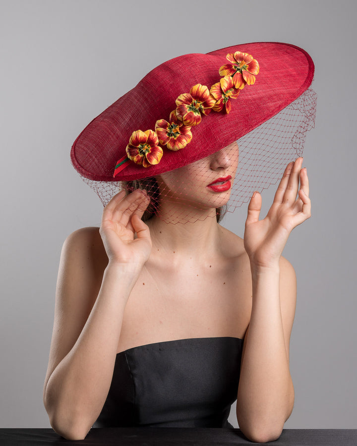 The female model is posing, wearing
Jericho, the deep red sinamay wide-brim fascinator with a sculptural pointed shape, decorated with orange and burgundy pansy flowers and a matching red birdcage veil. Designed and made by Merve Bayindir Millinery.
