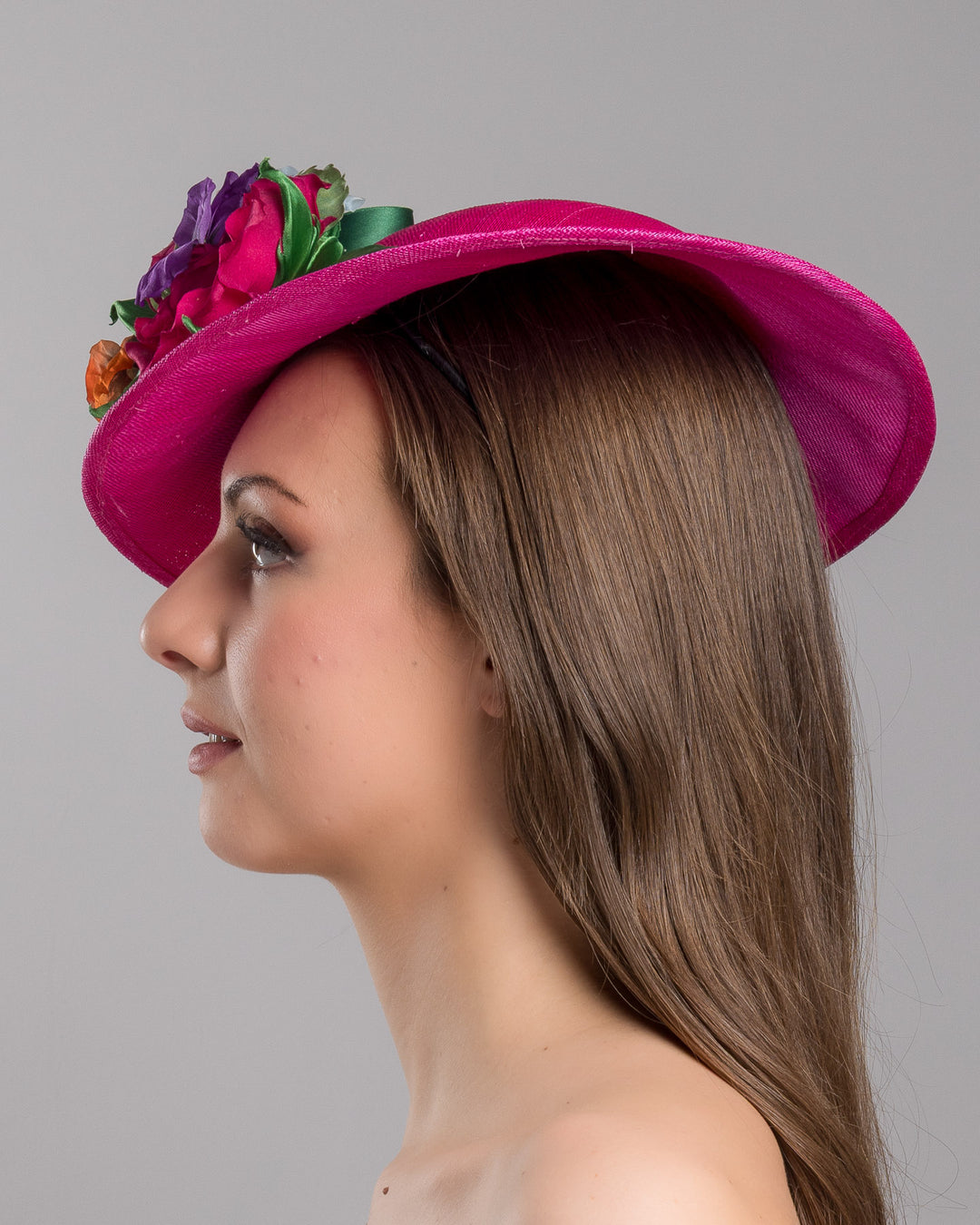 The model is showcasing the left side close up of the medium sized, hot pink medium teardrop saucer decorated with a bouquet of eye-catching pink roses, multi-coloured violets and emerald leaves. Designed and made by Merve Bayindir Millinery.

