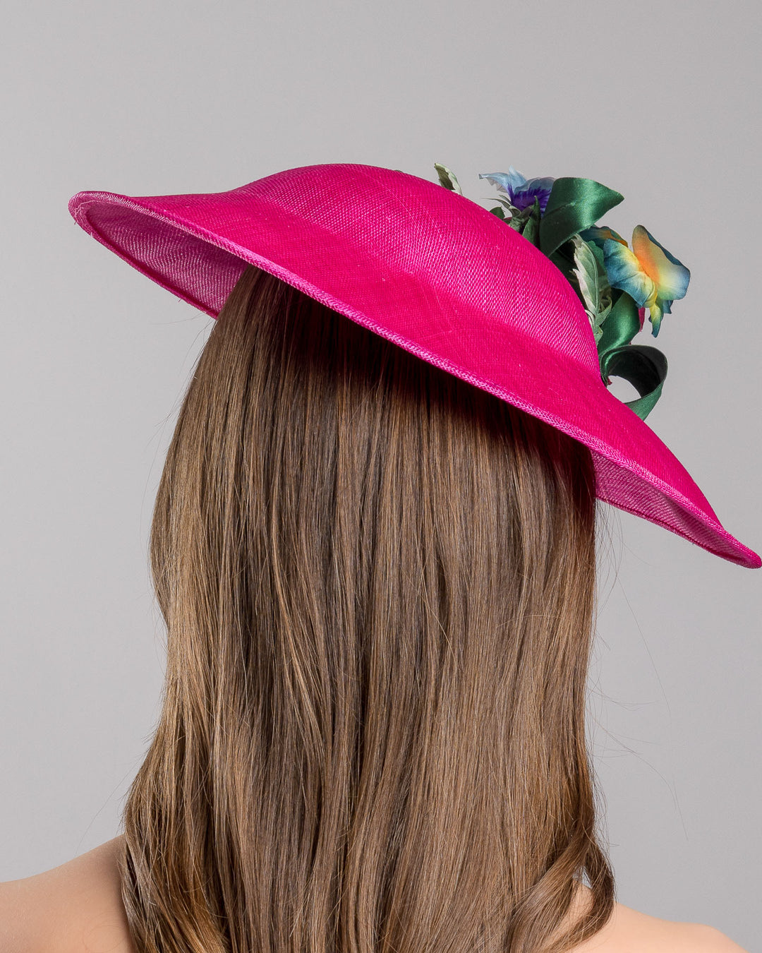 The model is showcasing the back side close up of the medium sized, hot pink medium teardrop saucer decorated with a bouquet of eye-catching pink roses, multi-coloured violets and emerald leaves. Designed and made by Merve Bayindir Millinery.
