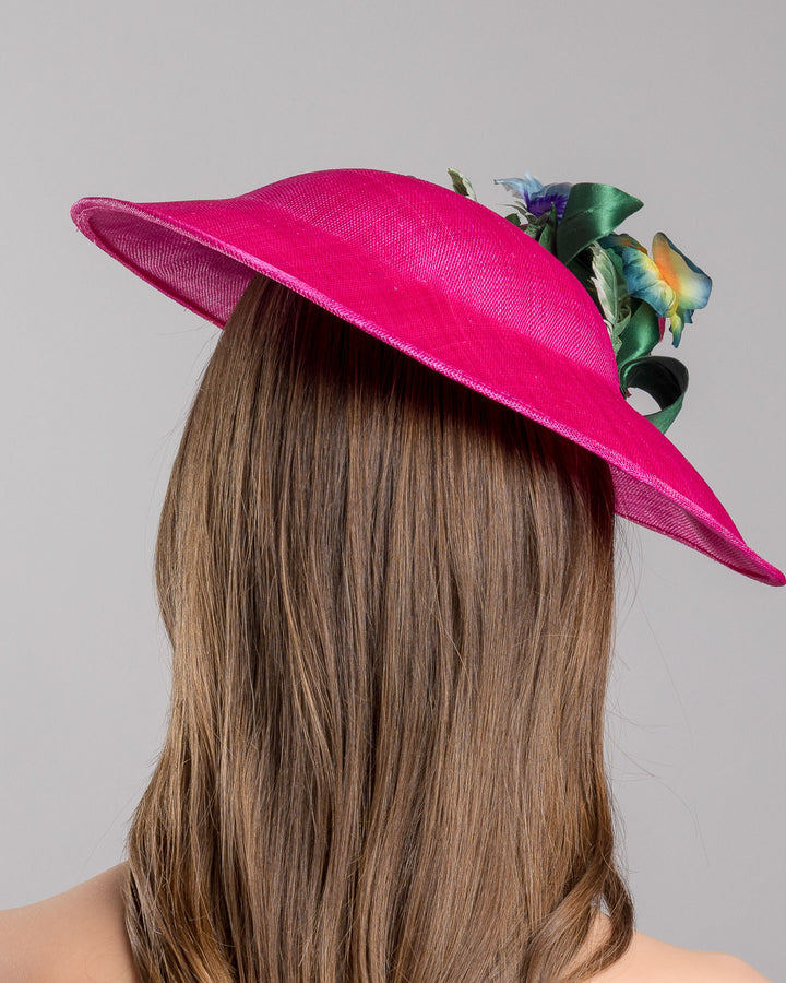 The model is showcasing the back side close up of the medium sized, hot pink medium teardrop saucer decorated with a bouquet of eye-catching pink roses, multi-coloured violets and emerald leaves. Designed and made by Merve Bayindir Millinery.
