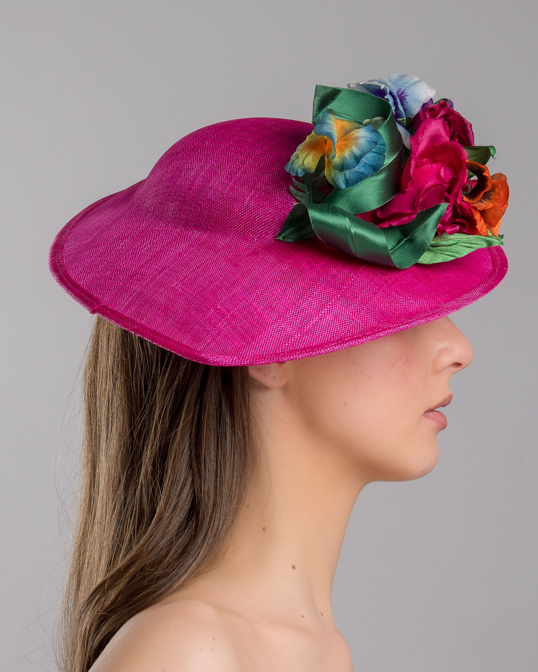 The model is showcasing the right side close up of the medium sized, hot pink medium teardrop saucer decorated with a bouquet of eye-catching pink roses, multi-coloured violets and emerald leaves. Designed and made by Merve Bayindir Millinery.
