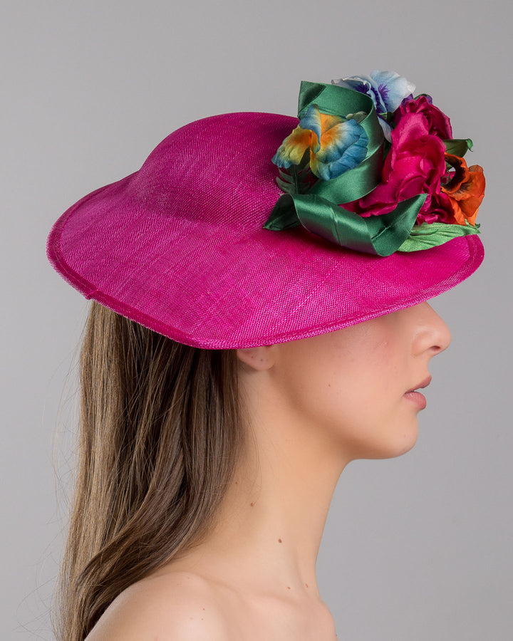 The model is showcasing the right side close up of the medium sized, hot pink medium teardrop saucer decorated with a bouquet of eye-catching pink roses, multi-coloured violets and emerald leaves. Designed and made by Merve Bayindir Millinery.

