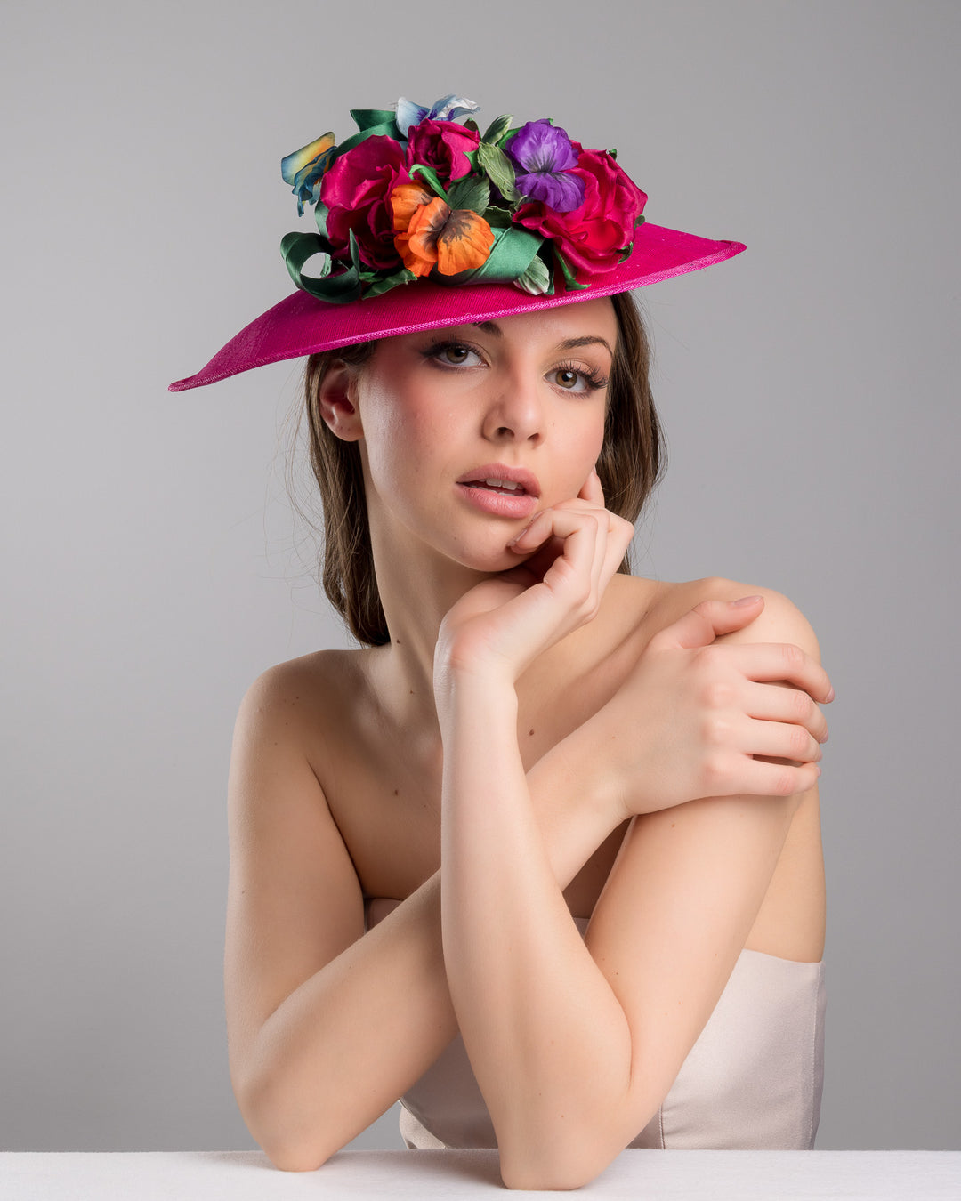 The female model is posing, wearing Jalen the medium sized, hot pink medium teardrop saucer decorated with a bouquet of eye-catching pink roses, multi-coloured violets and emerald leaves. Designed and made by Merve Bayindir Millinery.
