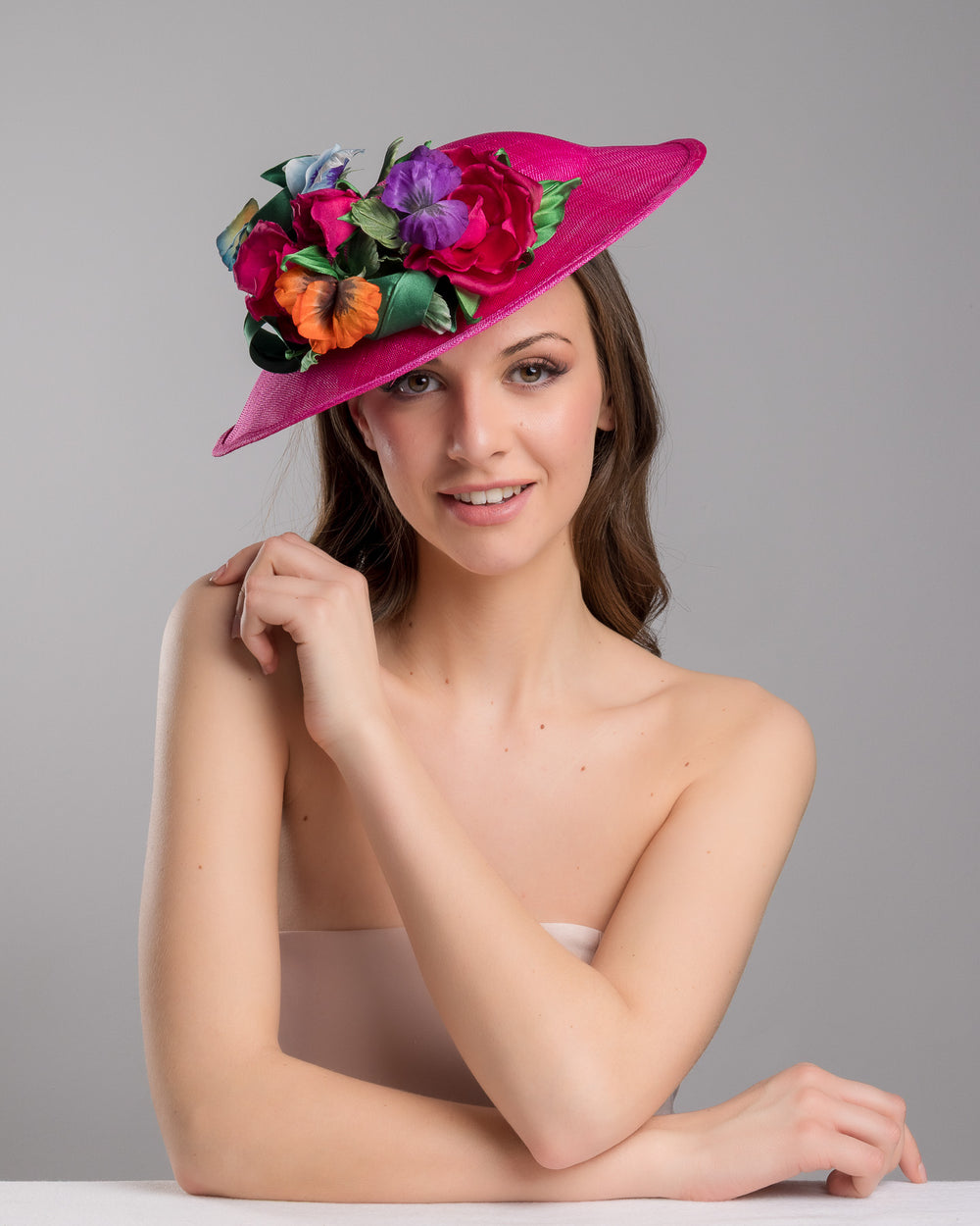 The female model is posing, wearing Jalen the medium sized, hot pink medium teardrop saucer decorated with a bouquet of eye-catching pink roses, multi-coloured violets and emerald leaves. Designed and made by Merve Bayindir Millinery.
