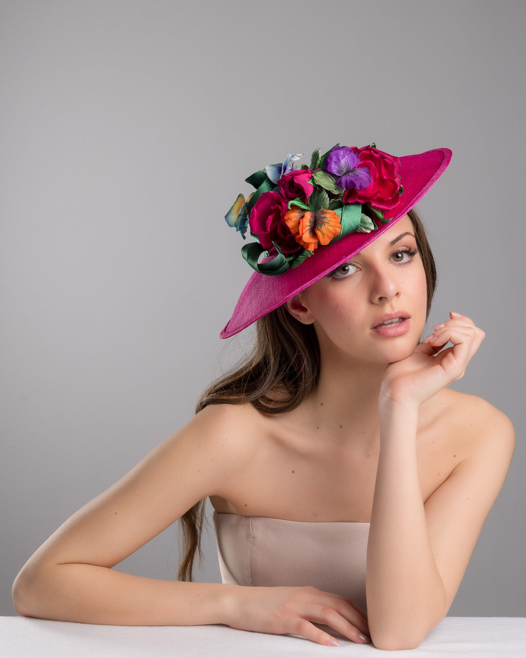The female model is posing, wearing Jalen the medium sized, hot pink medium teardrop saucer decorated with a bouquet of eye-catching pink roses, multi-coloured violets and emerald leaves. Designed and made by Merve Bayindir Millinery.
